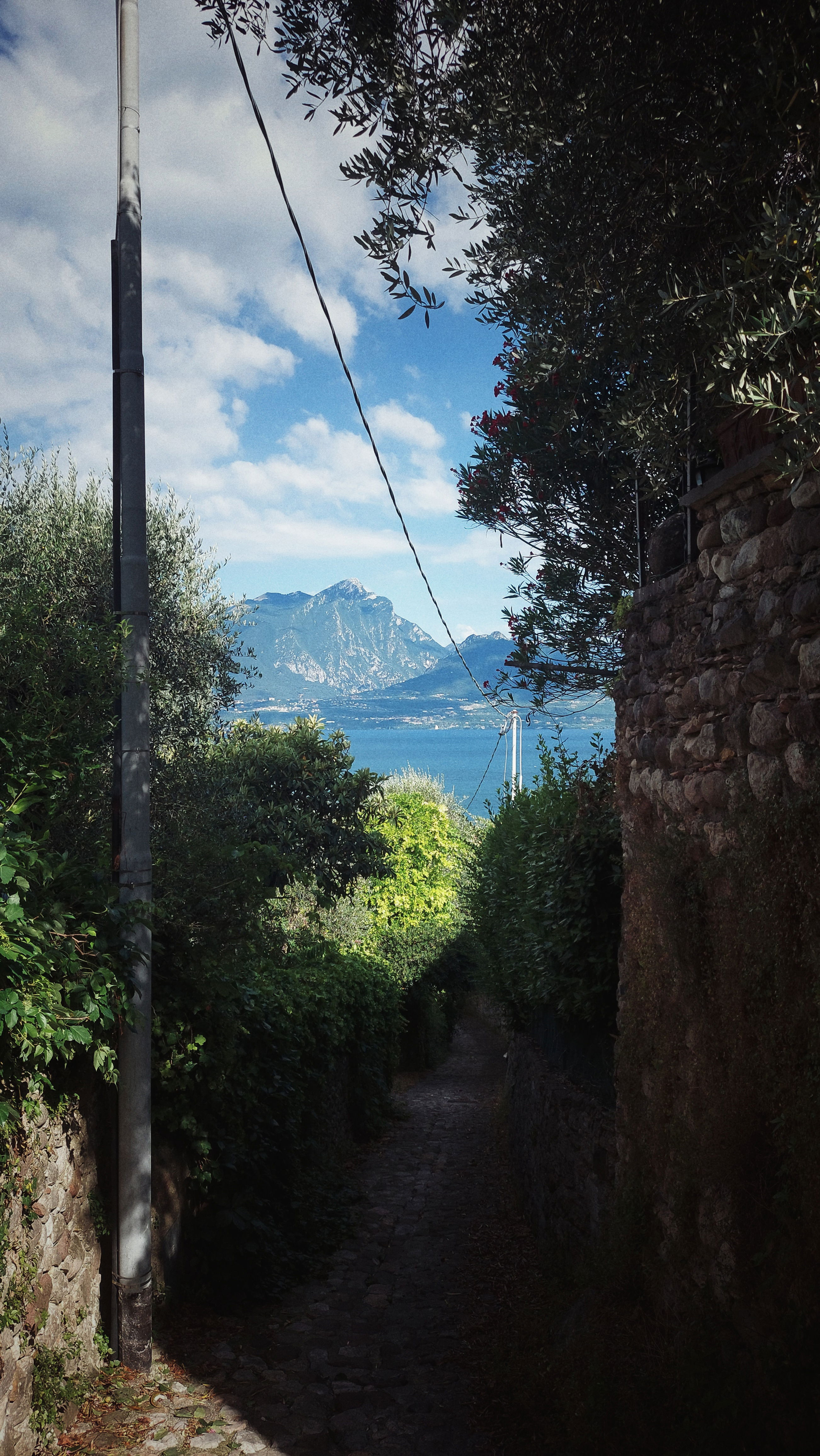 Looking down a narrow walk. Trees, old walls, mountains and a lake in the background.