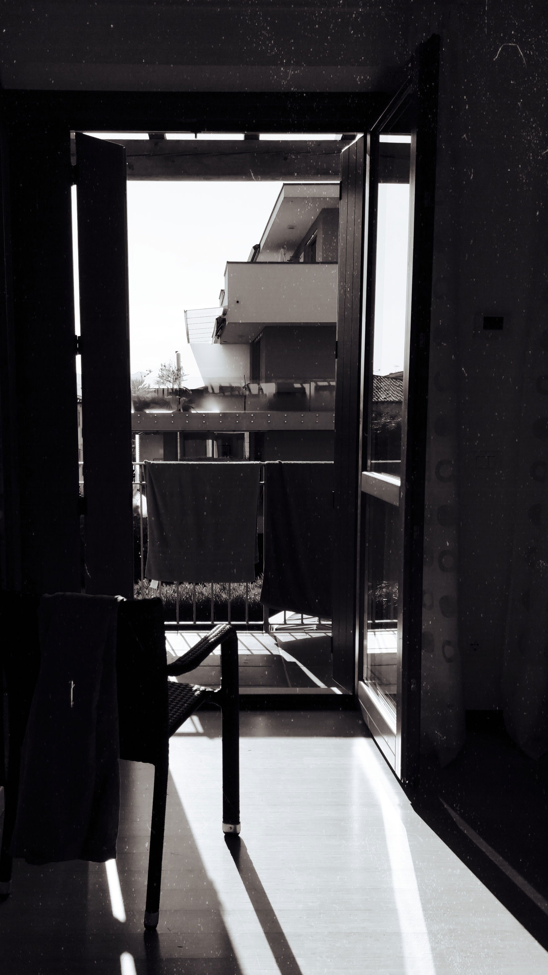 An open terrace window. A chair. A contemporary building outside. Strong black/white contrast.