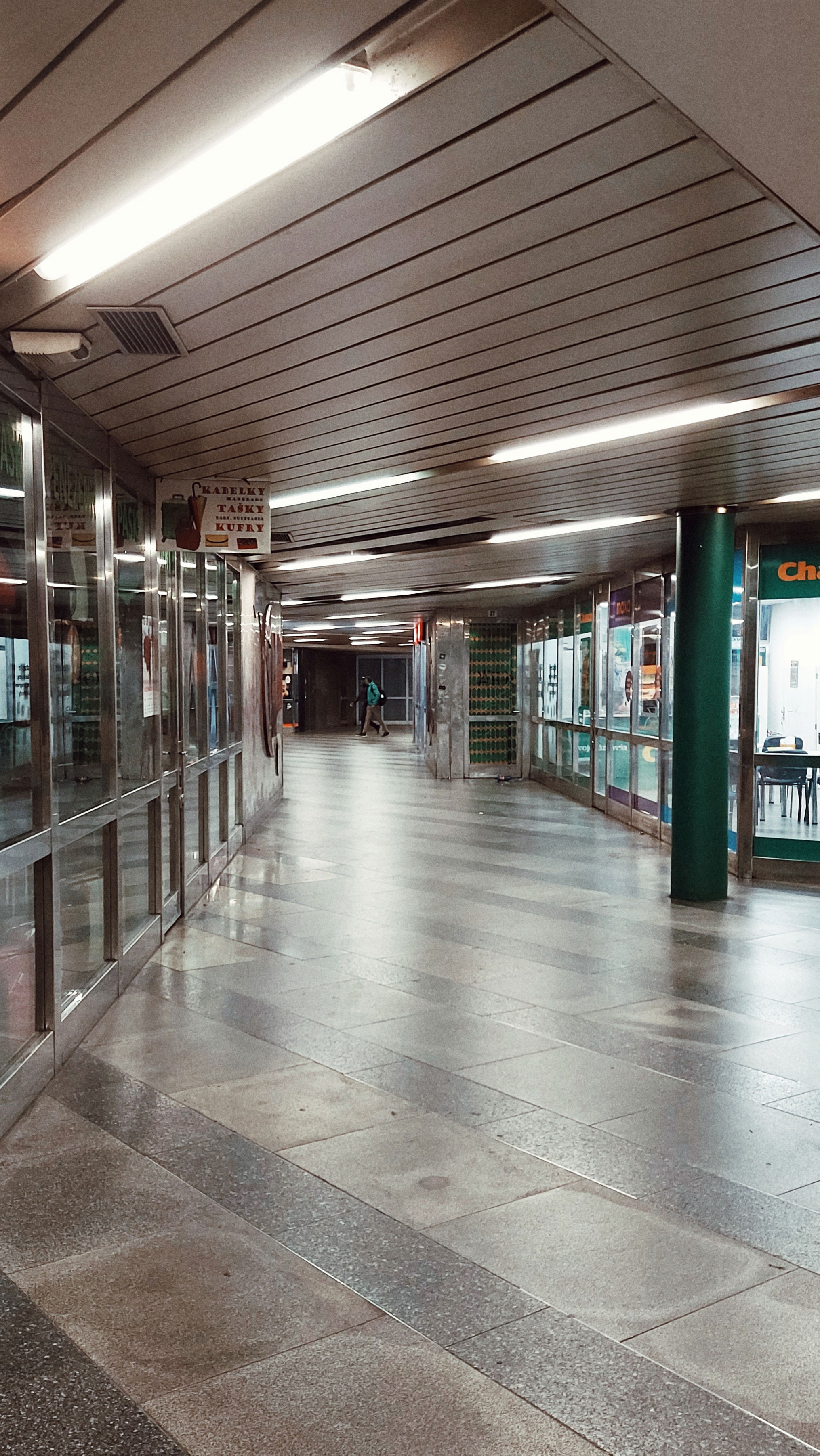 Lights and reflecting floor tiles in an empty metro station. Two stores left and right behind glass walls, the right one lit but also mostly empty.