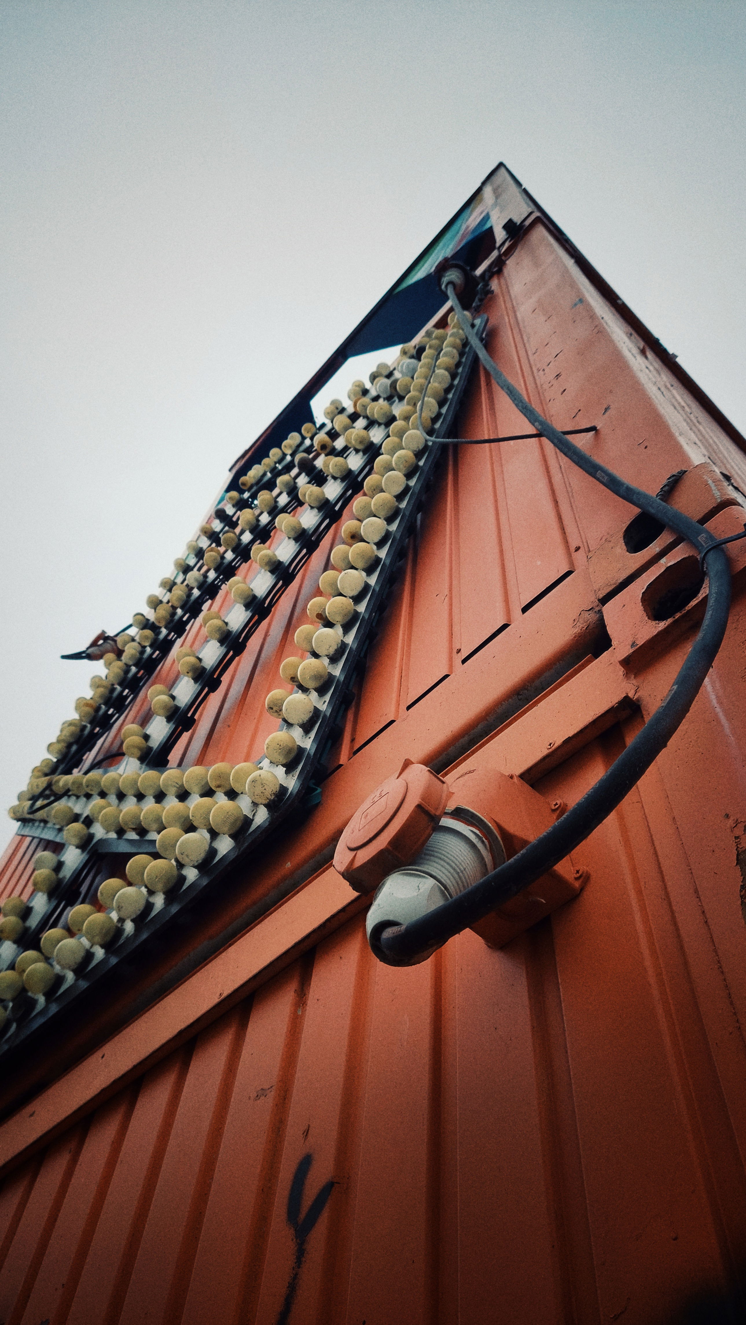A wall of red metal containers in front of white sky. There's a structure of light bulbs on the left part, and a thick wire connected to a power socket in front. 