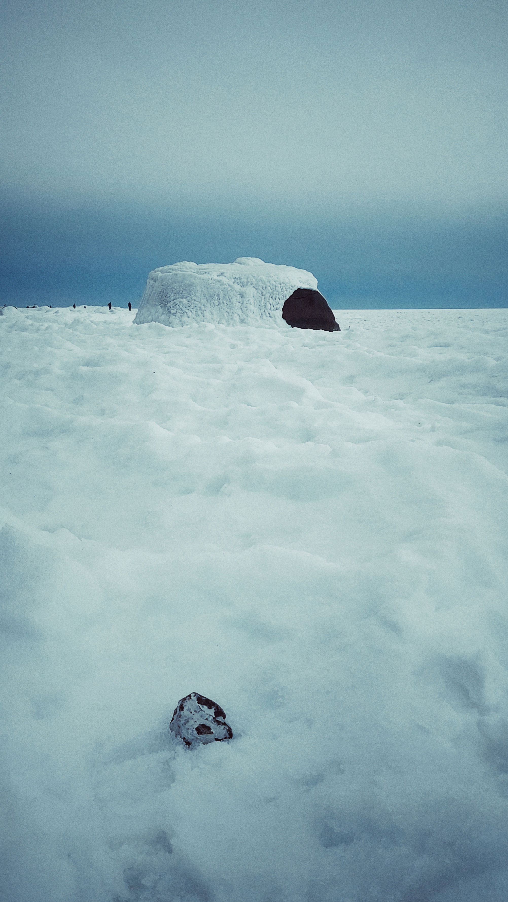 A small stone resting in the snow, a larger rock in the back covered by ice.