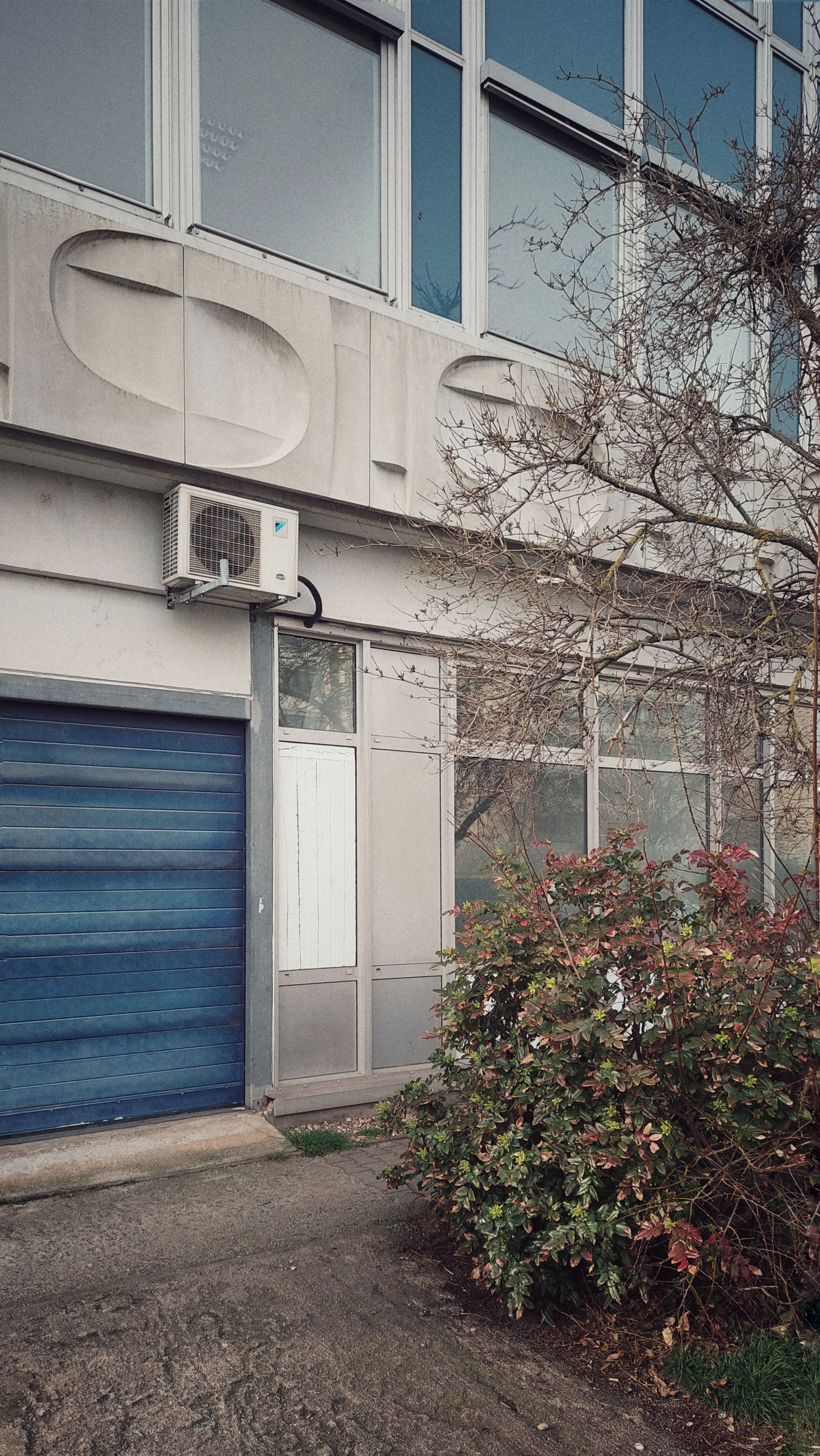 An East Modernist facade, concrete, glass, a blue sheet metal door on the left, blue elements in the window front right. Bushes hiding parts of the building.