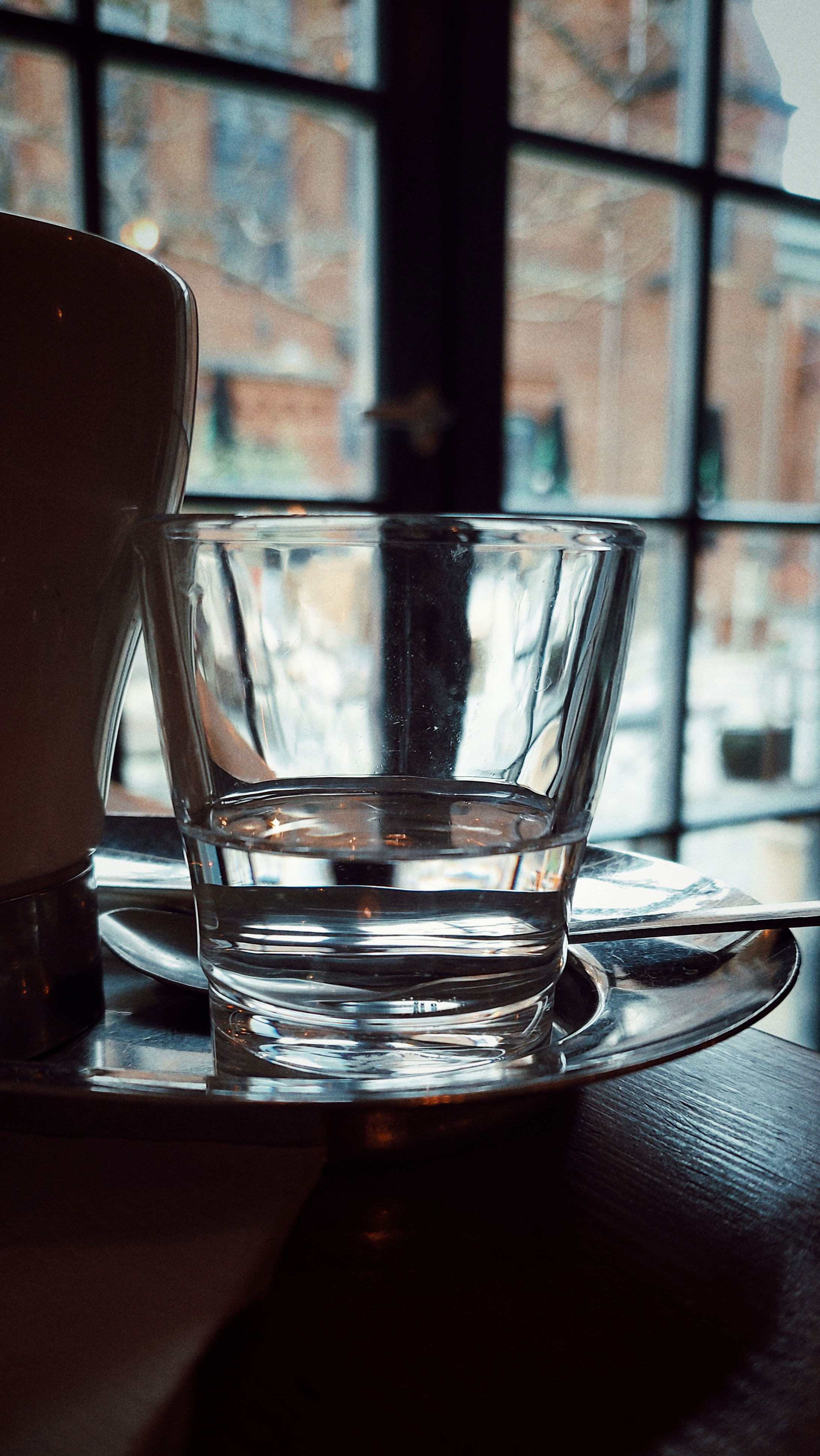 A glass of water near a coffee mug, closeup. Windows and a building structure in the back. 