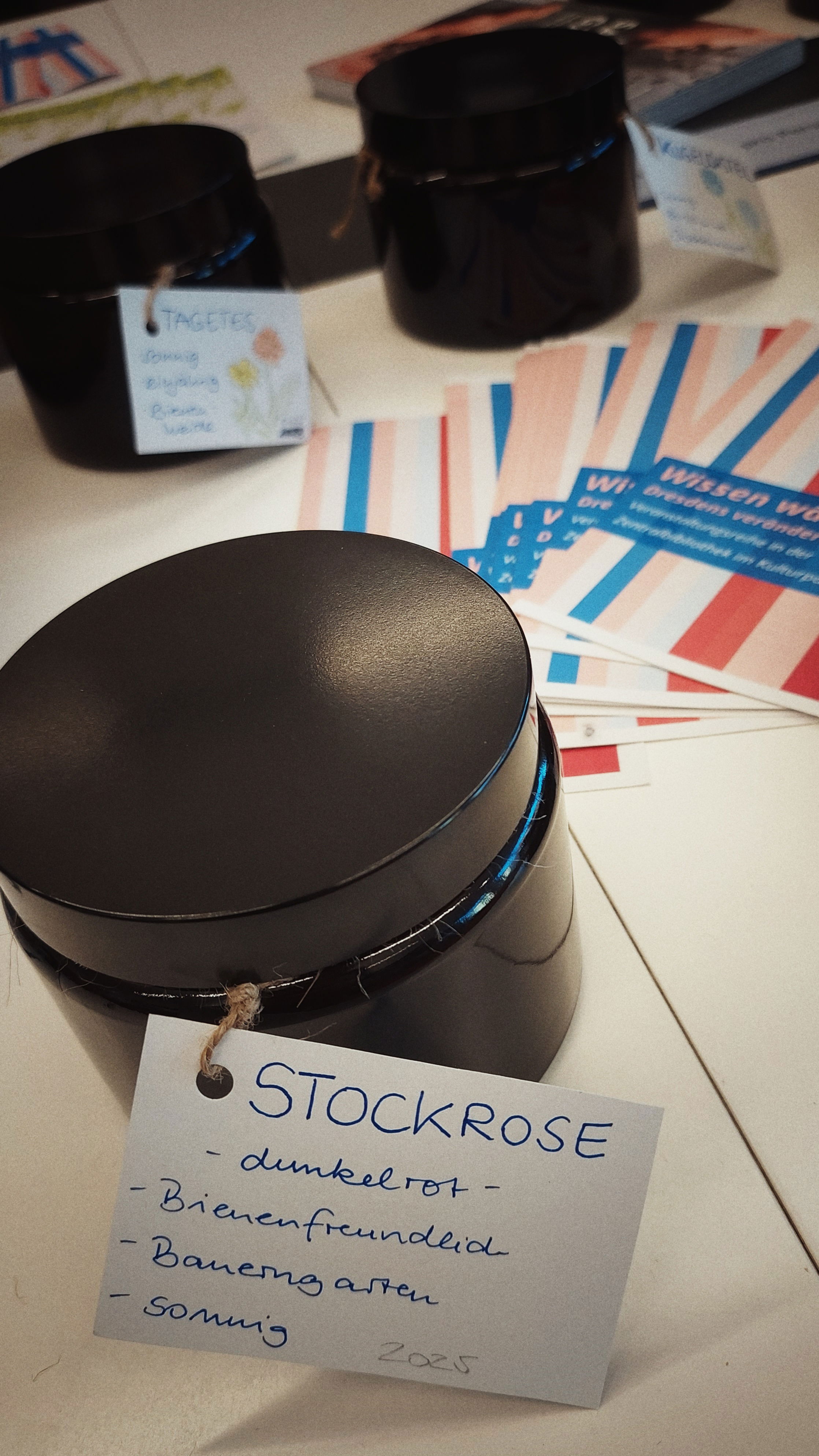 Three black round boxes on a table, each of them labeled, the front one label reading Stockrose, dunkelrot, bienenfreundlich, Bauerngarten. Seeds for a certain kind of roses apparently. Some flyers, red, blue, white color, in between.