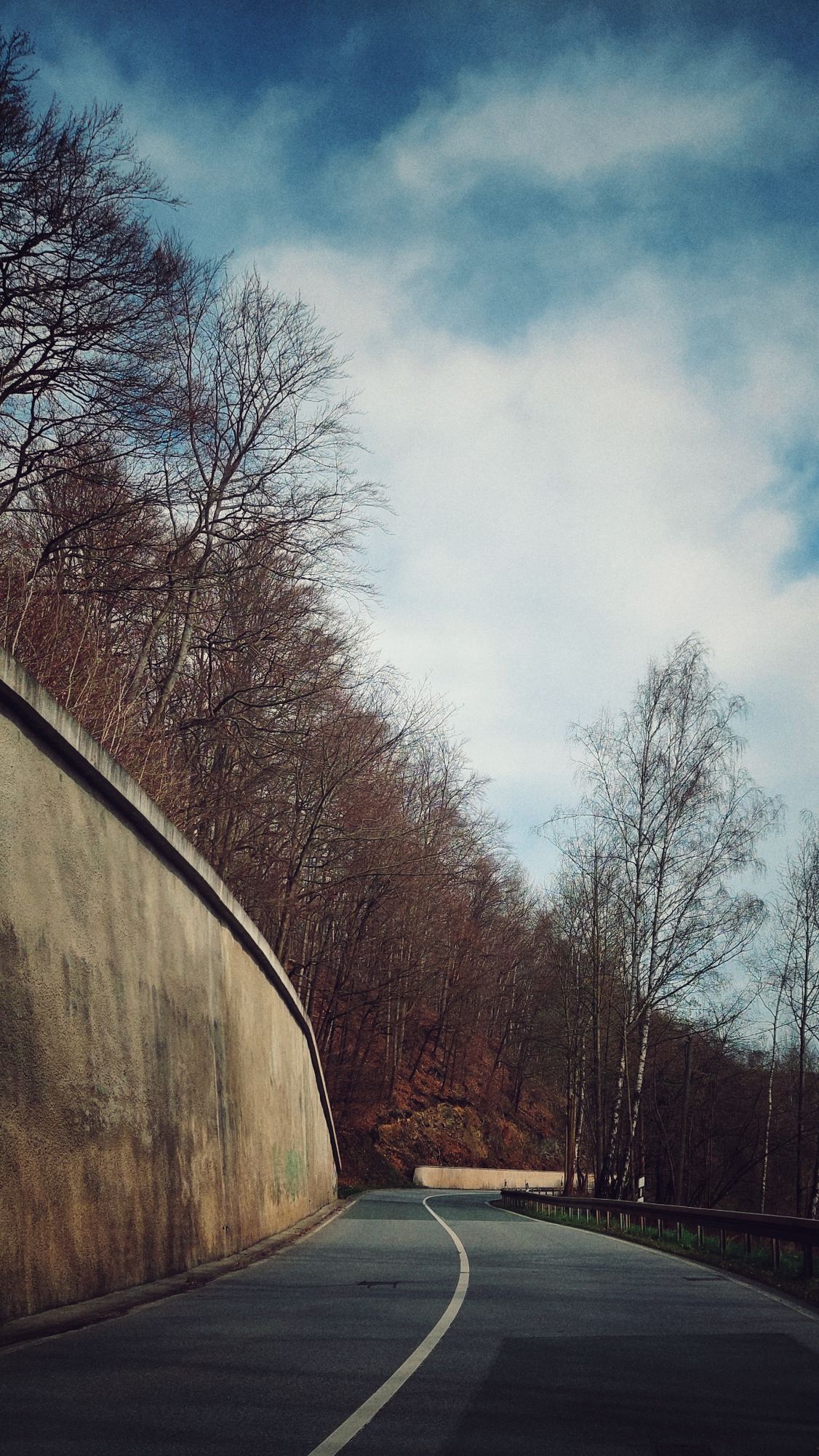 A street constrained by a strong wall. Hills, forest behind, a cloudy blue sky above. 