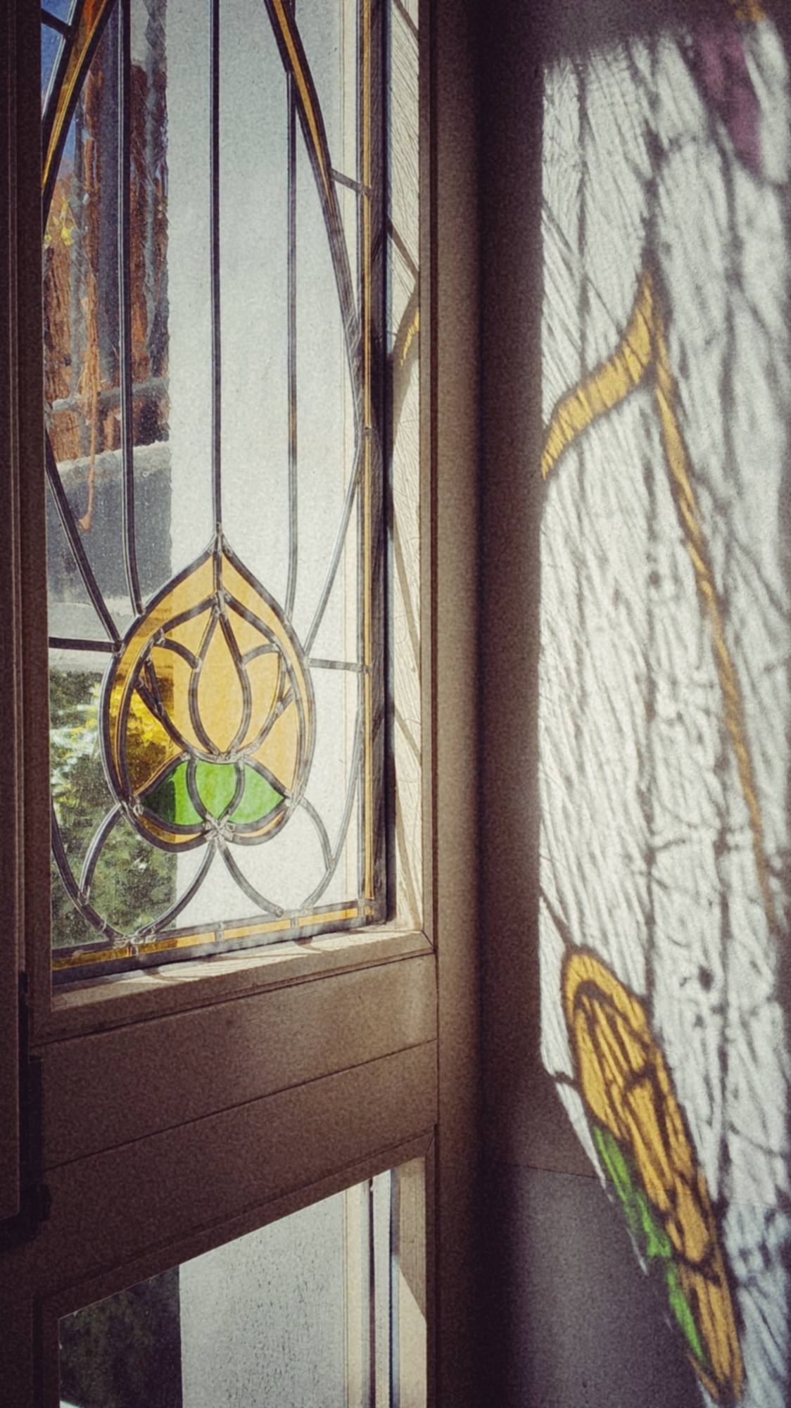 Light through an old window in a staircase. Tgs window holds an ornament made of green and yellow glass depicting an abstract flower in a structure of black ellipses and lines and the corresponding shadows are visible right of the window on an otherwise white wall.