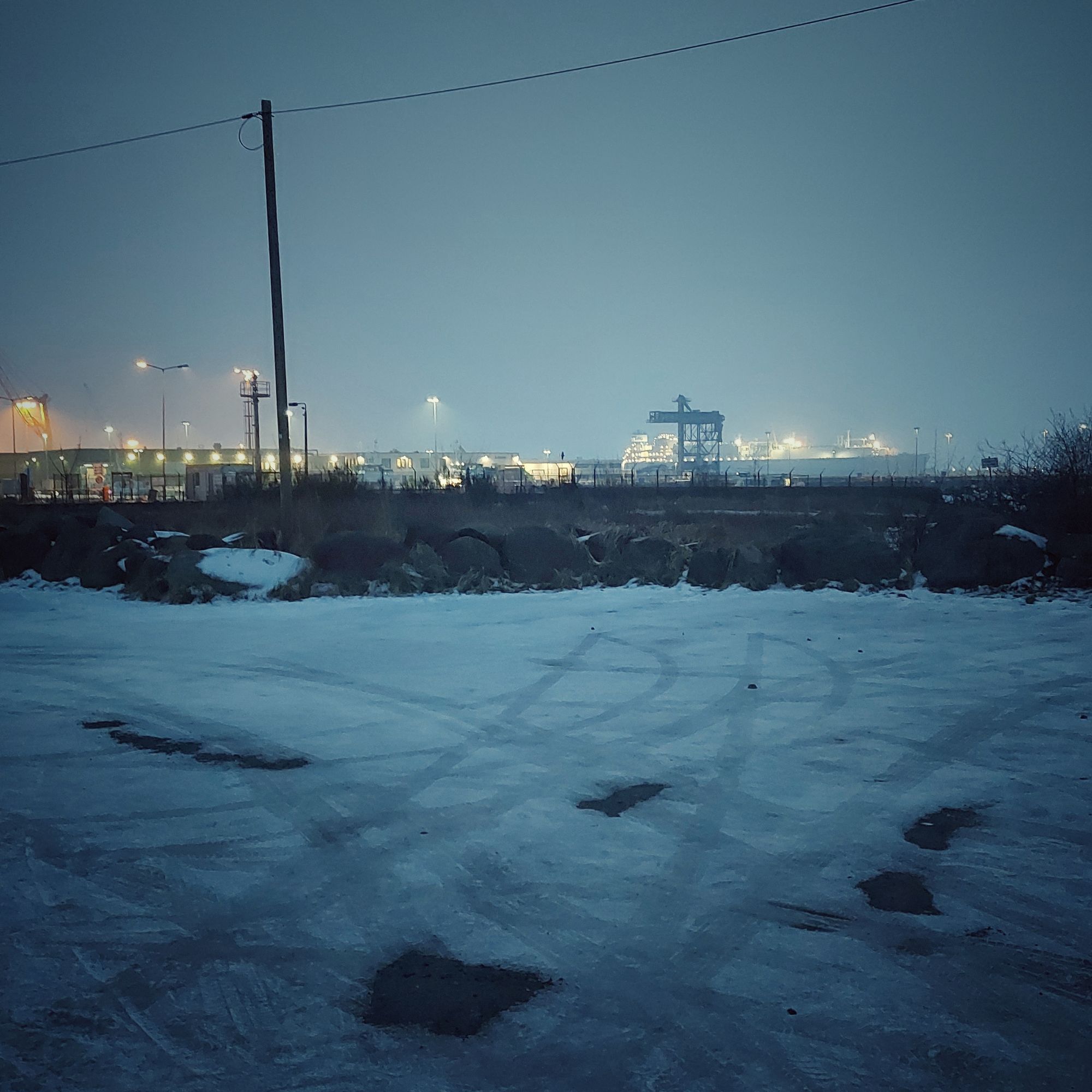 An icy parking area with car trails. A ferry terminal with a huge ship illuminated in the back.