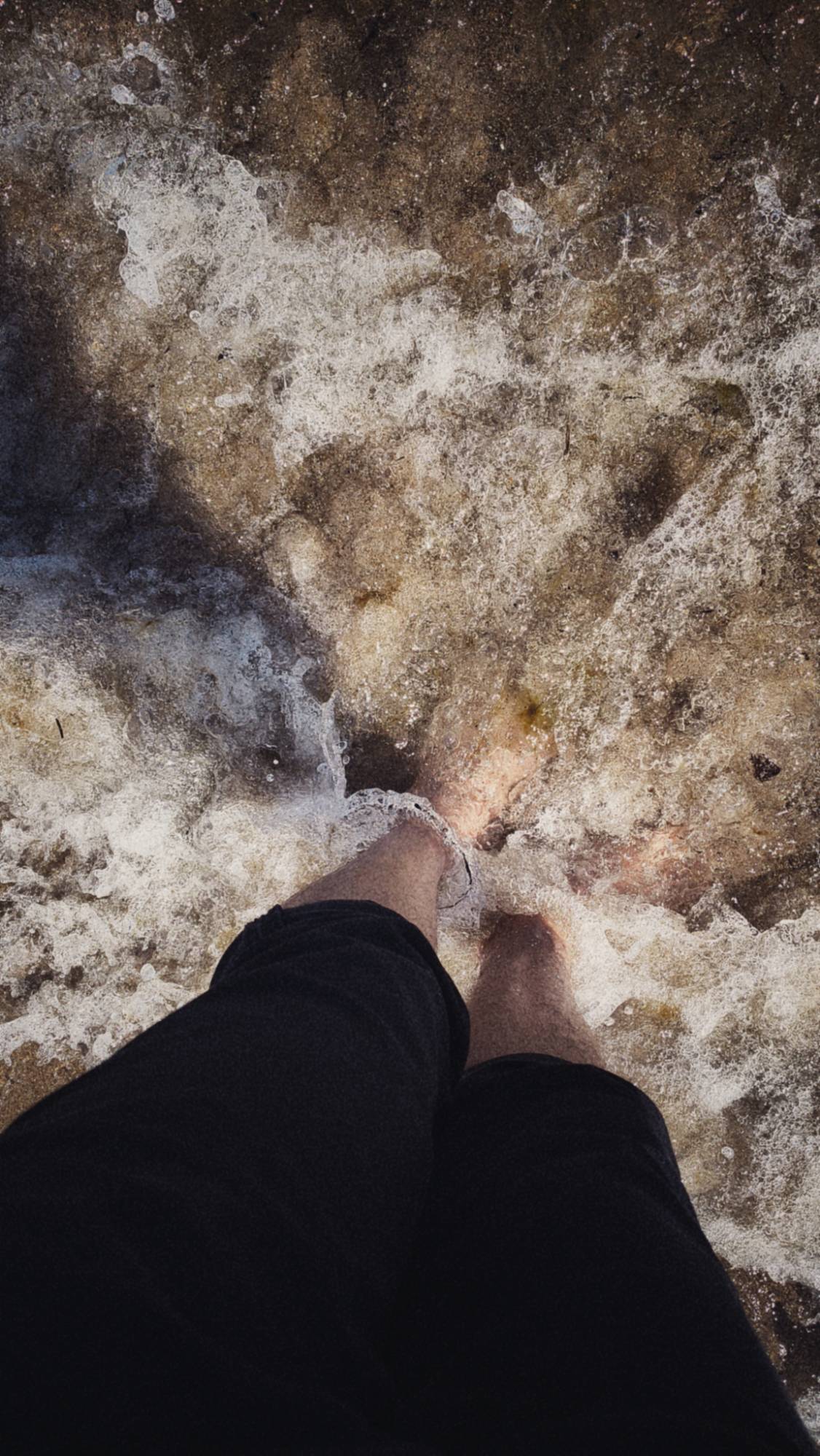 Legs with black trousers rolled up, naked feet in waves.